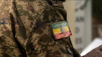 L'image montre un homme portant un uniforme militaire camouflé. Sur la manche de son uniforme, il y a un écusson tricolore représentant le drapeau éthiopien. Ce drapeau est composé de trois bandes horizontales : la bande supérieure est verte, la bande du milieu est jaune et la bande inférieure est rouge. Au centre, on retrouve une étoile bleue entourée d'un cercle doré. L'ensemble de la scène donne une impression de force et de discipline militaire.