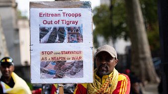 L'image montre un homme qui tient une pancarte haute, sur laquelle sont affichées des images et des textes en anglais. La pancarte exprime des revendications concernant le retrait des troupes érythréennes de la région du Tigré, en Éthiopie. Les mots en gras, "Eritrean Troops Out of Tigray" et "STOP USING STARVATION AS A WEAPON OF WAR", soulignent un appel urgent pour mettre fin à la souffrance causée par la guerre. L'homme porte un chapeau gris et est vêtu d'une écharpe colorée, probablement aux couleurs de l'Éthiopie, ce qui suggère un engagement émotionnel et une participation à une manifestation. En arrière-plan, on peut apercevoir d'autres personnes qui semblent partager des préoccupations similaires. L'ambiance est chargée d'émotion et de détermination.