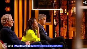 L'image montre un plateau de télévision lors d'une émission intitulée "France-Afrique : Rupture ou nouveau départ ?". Trois personnes sont assises sur un canapé, éclairées par des lumières chaudes. À gauche, un homme avec des lunettes et des cheveux grisonnants porte un pull sombre. Au centre, une femme, habillée d'un pull lumineux, semble attentive et réfléchie. À droite, un homme aux cheveux courts et argentés affiche une expression sérieuse. En arrière-plan, on peut apercevoir des fenêtres illuminées qui donnent une ambiance chaleureuse à l'ensemble. Le décor est moderne, avec des éléments graphiques en arrière-plan qui ajoutent à l'esthétique de l'émission.