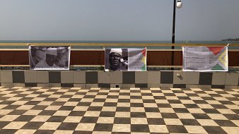 L'image montre un espace extérieur avec un sol en carreaux de damier, alternant entre des teintes claires et foncées. En arrière-plan, on aperçoit une étendue d'eau qui pourrait être la mer ou un lac, sous un ciel clair. Sur un muret, plusieurs affiches sont accrochées. Ces affiches semblent contenir du texte et des images en rapport avec un événement ou une exposition. L'environnement dégage une atmosphère calme, avec un éclairage naturel et une vue agréable sur l'eau.