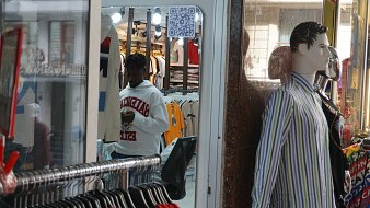 Dans cette image, nous voyons un magasin de vêtements. À l'avant, il y a un mannequin habillé d'une chemise rayée aux couleurs claires, affiché sur une vitrine. Derrière le mannequin, il y a un homme, qui porte un sweat à capuche blanc et semble utiliser son téléphone. Le magasin est bien éclairé, avec des étagères remplies de t-shirts colorés visibles à l'intérieur. Le fond de l'image montre une ambiance animée et des vêtements suspendus.