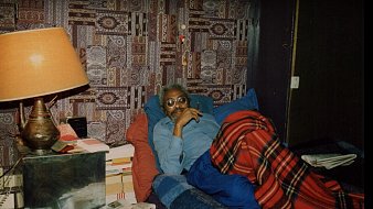 L'image montre un homme allongé sur un lit, recouvert d'une couverture à carreaux rouges et noirs. Il est vêtu d'une chemise bleue et porte des lunettes. L'homme semble pensif, avec un léger sourire. En arrière-plan, on distingue un mur tapissé d'un motif coloré et complexe. À côté du lit, une lampe de table en métal émet une lumière douce. Il y a également des piles de livres et de journaux sur une table à proximité, ce qui donne une impression de confort et d'intimité dans la pièce.