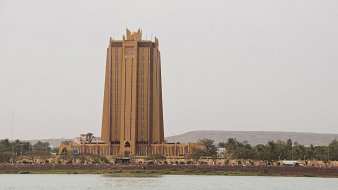 L'image montre un bâtiment imposant qui semble être une tour, située près de l'eau. Ce bâtiment, d'une teinte dorée, a une architecture moderne et élancée. Sa structure se soulève vers le ciel, avec des lignes verticales marquées et des détails décoratifs au sommet, donnant une impression de grandeur. En arrière-plan, on aperçoit quelques collines et des arbres, tandis qu'au premier plan, on voit la surface calme de l'eau. Le ciel est légèrement nuageux, créant une ambiance tranquille et légèrement brumeuse autour de cette scène urbaine.
