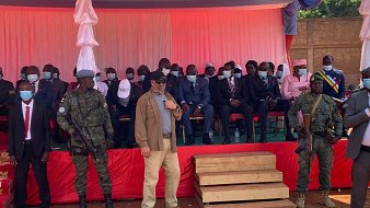 L'image montre une scène d'événement en plein air, probablement une cérémonie officielle. Au premier plan, deux soldats en uniforme, l'un avec un casque et l'autre portant une veste militaire, se tiennent debout à côté de deux marches en bois. Derrière eux, un groupe de personnes est assis sur des chaises, en grande partie habillées de manière formelle, certaines portant des masques. Les hommes et les femmes sont alignés en rang, sur un fond de tissu rouge et blanc. L'ambiance semble solennelle, avec des drapeaux et des décorations typiques des événements institutionnels. Le décor est éclairé par une lumière naturelle, et l'atmosphère pourrait évoquer une cérémonie de discours ou une célébration publique.