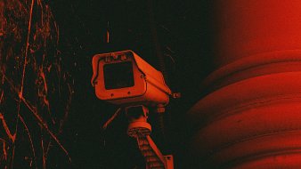 L'image représente une caméra de surveillance fixée à un mur. La caméra, avec son boîtier rectangulaire et son objectif, est peinte en gris clair. Elle est montée sur un support qui lui permet de pivoter, capturant ainsi une vaste zone de surveillance. L'arrière-plan est d'un rouge intense, créant une atmosphère sombre et légèrement inquiétante. Des textures sombres et des motifs abstraits se dessinent autour, ajoutant à l'effet dramatique de l'image. Cette combinaison de couleurs et de formes suggère une ambiance de vigilance et de sécurité.