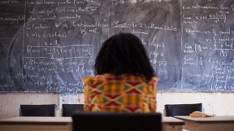 Dans cette image, on voit une personne assise, de dos, face à un grand tableau noir couvert d'écritures en craie blanche. Les formules mathématiques et les notes semblent complexes, suggérant un environnement académique ou d'apprentissage. La personne porte un vêtement coloré avec des motifs vibrants, ajoutant une touche de culture. L'ambiance est studieuse, et l'environnement évoque une salle de classe ou un espace de travail. Le contraste entre les couleurs vives du vêtement et la sombre surface du tableau crée une composition visuelle intéressante.