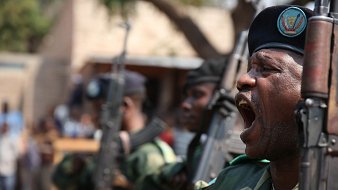 L'image montre un groupe de militaires en uniforme, en position de parade. L'un d'eux, au premier plan, est en train de crier ou de donner un commandement, affichant une expression de détermination. Ils portent des bérets sombres et tiennent des fusils en position verticale. En arrière-plan, on peut apercevoir d'autres soldats ainsi qu'un groupe de personnes qui semblent regarder la scène. L'environnement est extérieur, probablement lors d'un événement officiel ou d'une cérémonie. L'atmosphère est solennelle et dynamique, évoquant un sentiment de force et de discipline.