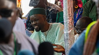 L'image montre un groupe de personnes rassemblées dans un environnement animé, probablement lors d'un événement public. Au premier plan, un homme souriant est vêtu d'un habit traditionnel, avec un turban ou une coiffe verte. Il semble engagé dans une discussion, ses mains gesticulant pour accompagner ses paroles. En arrière-plan, d'autres personnes sont présentes, certaines en uniforme, suggérant un contexte formel ou officiel. La scène est remplie de couleurs vives, et l'atmosphère semble dynamique et interactive.