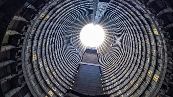 Imaginez vous tenir au centre d'une immense structure circulaire. La vue se dirige vers le haut, où un puits de lumière éclaire l'espace depuis l'ouverture au sommet. Les parois de ce bâtiment sont composées de béton et forment des motifs en spirale, créant une impression de profondeur et de hauteur. Les fenêtres qui bordent les étages, bien que nombreuses, laissent passer une lumière diffuse qui ajoute une sensation d'apaisement. Le sol est plat et peut sembler rugueux, contrastant avec la douceur de la lumière qui découpe le vide en haut. L'atmosphère est à la fois vide et pleine de possibilité, un mélange d'architecture industrielle et de nature, le tout se rejoignant dans ce grand espace ouvert.