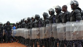 L'image montre une formation de policiers en tenue anti-émeute. Ils se tiennent en ligne, rigides et organisés, en portant des casques de protection et des gilets pare-balles. Chaque policier tient un bouclier transparent en face de lui, prêt à faire face à une situation potentielle. Le sol est en terre battue et l'environnement semble être à l'extérieur, avec quelques nuages visibles dans le ciel. Au loin, on peut apercevoir des personnes qui observaient la scène. L'atmosphère suggère une présence de sécurité renforcée.