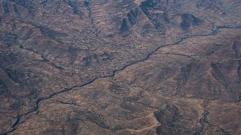 Cette image montre un paysage désertique vu du ciel. On peut observer de vastes étendues de terrain aride, striées par des vallées et des collines aux formes irrégulières. Au centre, un cours d'eau serpente à travers ce paysage, créant un contraste avec les zones sèches environnantes. Les couleurs dominantes sont des teintes de marron et de beige, évoquant un sol rocailleux et dénudé, tandis que la rivière apporte une touche de reflets plus sombres, suggérant des zones humides dans ce milieu. L'ensemble dégage une impression de vastitude et de sérénité, avec une nature sauvage encore peu altérée par l'homme.