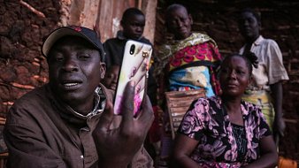 Cette image présente un groupe de personnes rassemblées à l'extérieur d'une maison. Au premier plan, un homme tient un téléphone portable et semble communiquer ou montrer quelque chose à ses interlocuteurs. Il porte une casquette et un manteau. À l'arrière-plan, plusieurs femmes regardent avec attention. Elles sont vêtues de vêtements colorés et affichent des expressions d'intérêt ou de curiosité. L'environnement est simple, avec des murs en terre crue et un éclairage qui suggère qu'il pourrait être tard dans la journée. L'atmosphère semble conviviale et axée sur la communication.