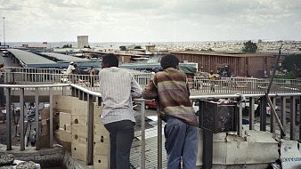 Dans cette image, on voit deux hommes debout sur un balcon surélevé, se penchant contre la balustrade. Ils regardent en bas un marché animé. Leurs silhouettes sont tournées vers l'extérieur, révélant un paysage urbain qui s'étend derrière eux, avec des bâtiments et des petites boutiques. L'environnement est vivant, rempli de personnes et d'activités. Le ciel est nuageux, ajoutant une ambiance mélancolique à la scène. Les vêtements des hommes suggèrent une certaine simplicité, avec des motifs discrets. L'ensemble dégage une atmosphère de contemplation et d'observation de la vie quotidienne en ville.