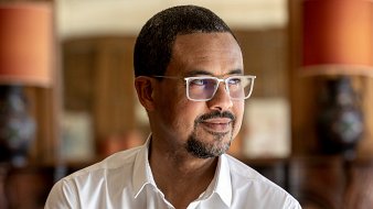 L'image présente un homme d'âge moyen, debout légèrement sur le côté. Il a la peau foncée et porte des lunettes avec une monture claire. Son expression est contemplative, avec un léger sourire aux lèvres. Il porte une chemise blanche, qui contraste avec son teint. En arrière-plan, on peut apercevoir des lampes avec des abat-jour orange et un décor en bois qui donne une ambiance chaleureuse et intime à l'ensemble. La lumière est douce, créant une atmosphère paisible.