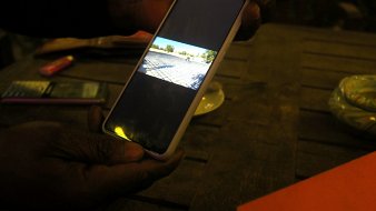 L'image montre une main tenant un téléphone portable, qui affiche une photographiée (ou une vidéo) d'un paysage. À l'arrière-plan, on peut imaginer une scène en extérieur, probablement un terrain ou une route. La lumière dans la pièce est tamisée, et il y a une table en bois visible. Sur la table, on peut apercevoir quelques objets, comme un petit paquet de cigarettes et un plateau. L'ambiance semble tranquille et décontractée, suggérant un moment de détente ou de partage.