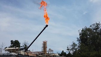 L'image présente une installation industrielle en plein air, probablement liée à l'extraction de pétrole ou de gaz. Au centre, un tube long et noir s'élève vers le ciel, d'où s'échappe une flamme vive et orange, créant un fort contraste avec le ciel bleu clair en arrière-plan. On aperçoit également des structures en métal, comme des réservoirs et des tuyaux, sur le site. De la végétation verte entoure l'installation, ajoutant une touche naturelle à cette scène industrielle. Le tout évoque une activité intense et potentiellement bruyante.