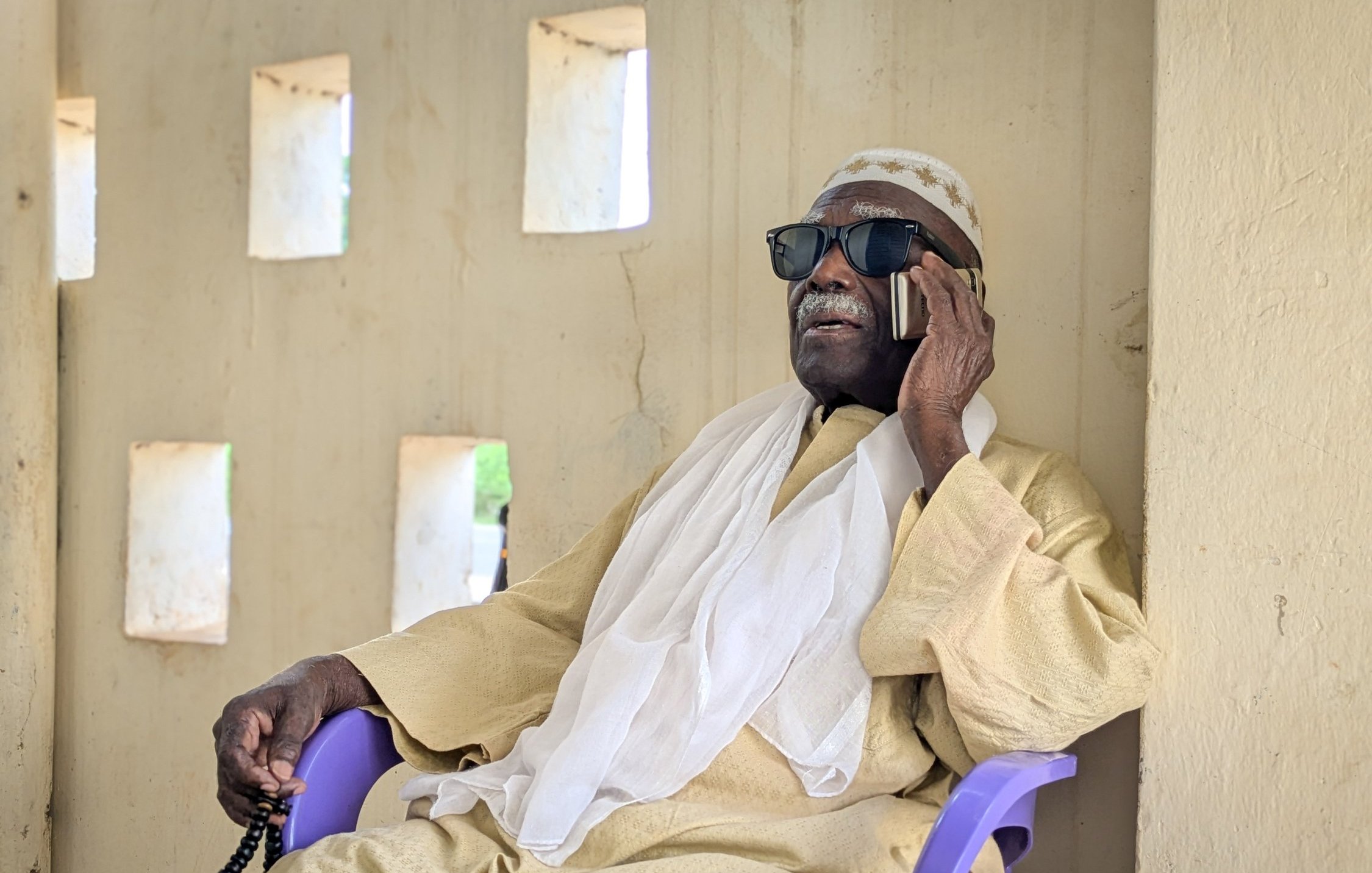 L'image montre un homme assis sur une chaise en plastique violette, contre un mur de couleur beige avec des fenêtres rectangulaires qui laissent passer une lumière douce. Il porte des lunettes de soleil noires et un vêtement traditionnel de couleur claire, accompagné d'un châle blanc drapé sur ses épaules. L'homme est en train de parler au téléphone, son visage exprime la concentration et la sérénité. L'environnement est calme, évoquant un moment de détente dans un cadre paisible.