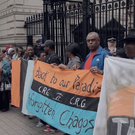 Dans cette image, un groupe de manifestants se rassemble devant un édifice imposant, probablement un bâtiment gouvernemental. Les personnes sont vêtues de vêtements variés, et certaines tiennent des pancartes. L'une des pancartes est colorée en orange, noir et bleu, et porte des inscriptions comme "Back to our Paradise" et "Forgotten Chagos". L'atmosphère est empreinte de détermination, avec des visages sérieux et engagés. Au fond, on peut voir des policiers surveillant la situation. L'environnement est urbain, avec des pavés au sol et des murs architecturaux ornés.