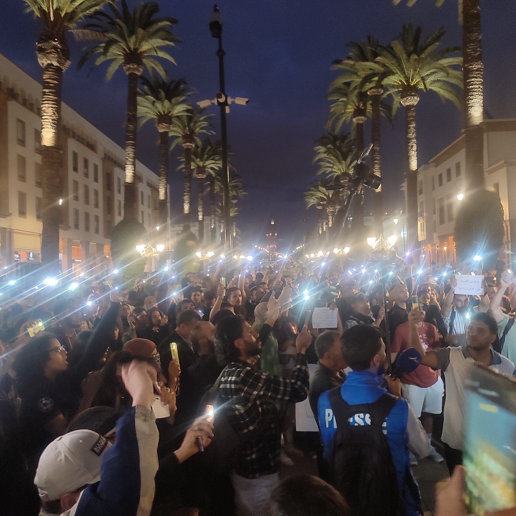 L'image montre une grande foule rassemblée dans une rue animée, éclairée par des lampadaires et entourée de palmiers. La scène se déroule en soirée, avec un ciel crépusculaire. Beaucoup de personnes tiennent des téléphones ou des lumières, créant un effet lumineux impressionnant. Les gens semblent mobilisés, certains tenant des pancartes et s'exprimant avec passion. L'atmosphère est énergique et engagée, reflétant une ambiance de manifestation ou d'événement public où les participants cherchent à faire entendre leur voix.