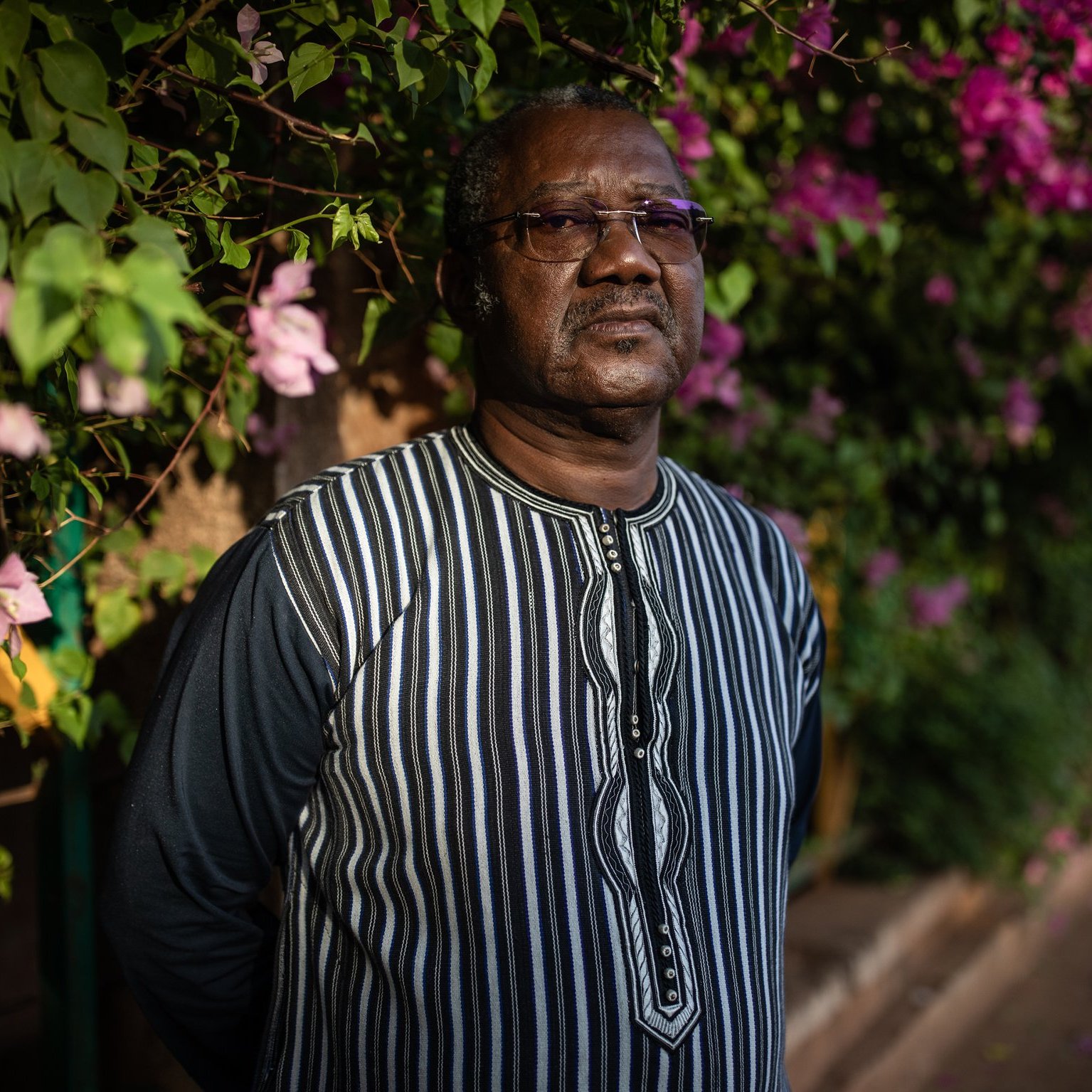 L'image montre un homme debout devant un mur recouvert de magnifiques bougainvilliers aux couleurs vives, principalement en rose et violet. Il porte une chemise à rayures noires et blanches, avec un col qui attire l'attention. Son expression est sérieuse et réfléchie, et il semble absorbé par ses pensées. En arrière-plan, on peut percevoir une ambiance douce et chaleureuse grâce à la lumière naturelle qui illumine la scène, accentuant la beauté des fleurs.