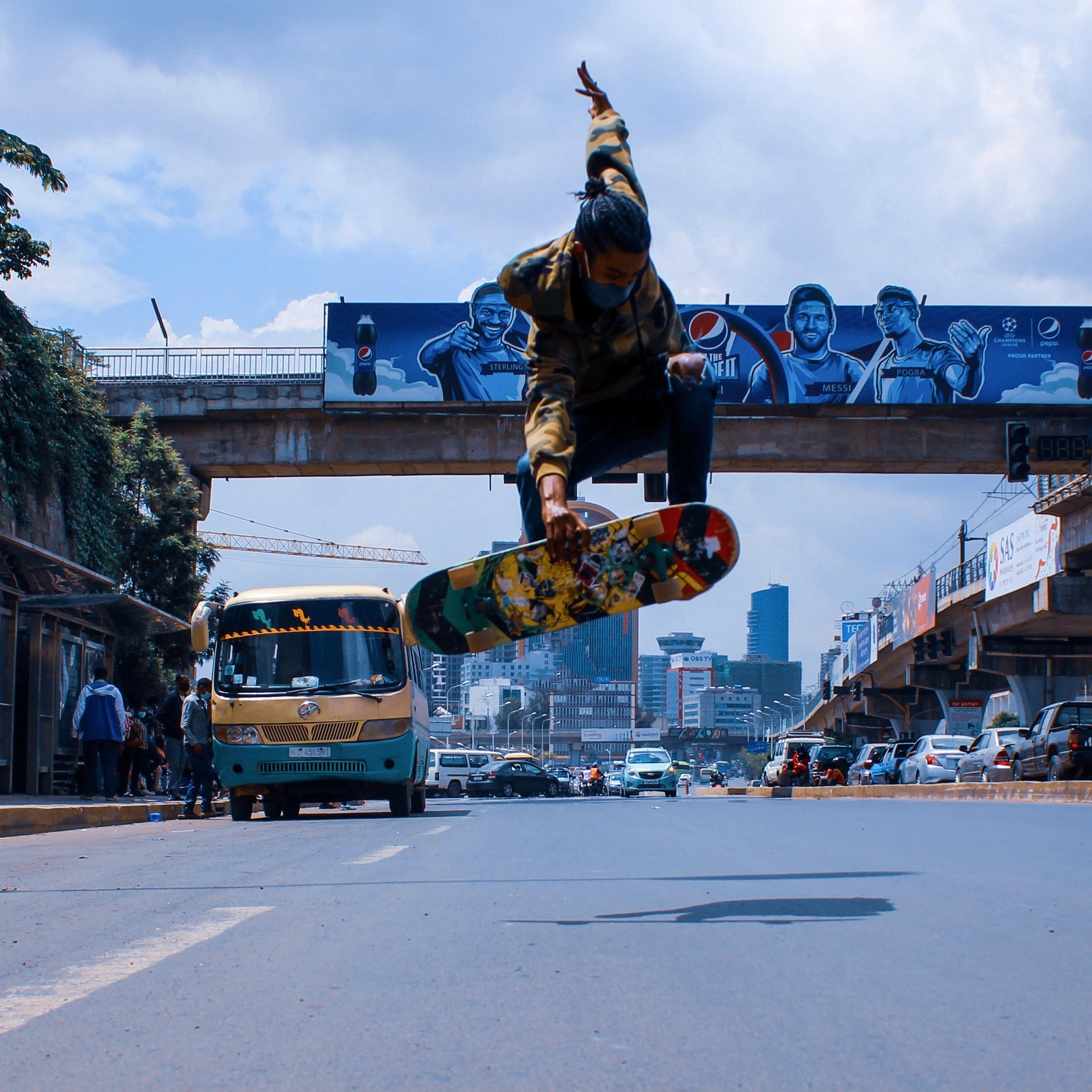 L'image montre un skateur en plein saut au-dessus de la route. Il est habillé d'un sweat à capuche avec des motifs et porte un skateboard coloré. En arrière-plan, il y a des bâtiments urbains, des graffitis sur un mur, et un bus qui passe, typique des grandes villes. La scène capture une ambiance dynamique et énergique, avec un ciel partiellement nuageux qui ajoute à l'atmosphère urbaine. Les paysages urbains et la présence d'autres personnes montrent l'animation de la ville.