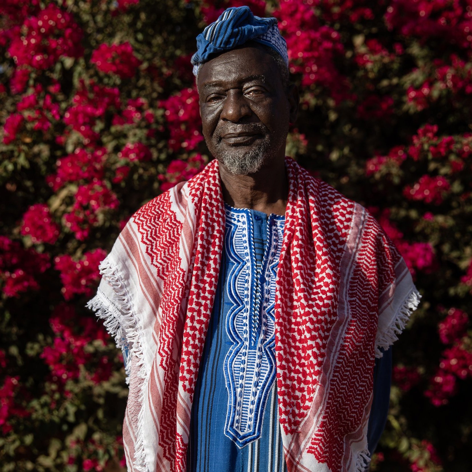 L'image montre un homme debout devant un fond vibrant de fleurs roses, probablement des bougainvilliers. Il porte une tenue traditionnelle riche en couleurs, avec un tissu bleu et des motifs complexes. Sur ses épaules, il a une écharpe ou un châle rouge et blanc, typique de certaines cultures. Son visage exprime sagesse et dignité, avec des traits marqués par le temps. Le contraste entre ses vêtements et les fleurs éclatantes crée une atmosphère chaleureuse et accueillante.