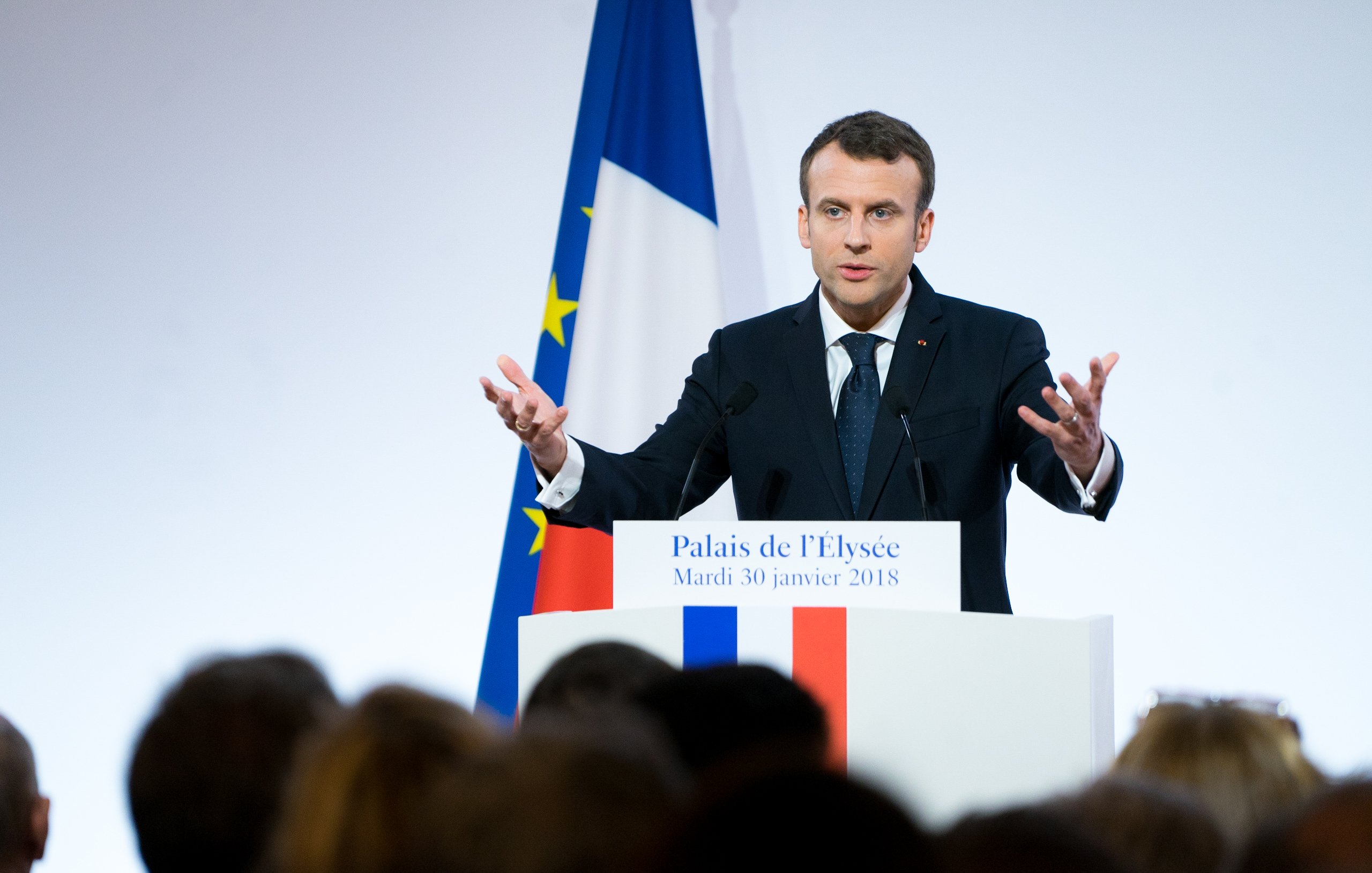 L'image montre un homme debout devant un podium, probablement en train de donner un discours. Il porte un costume sombre avec une cravate, et ses mains sont levées, ce qui indique qu'il met l'accent sur ses propos. En arrière-plan, on aperçoit le drapeau français aux couleurs bleu, blanc et rouge, ainsi qu'une autre bannière européenne. La scène semble se dérouler dans un cadre officiel, peut-être à l'Élysée, avec un public flou et attentif. L'atmosphère est sérieuse et engageante, marqué par le dynamisme du orateur.