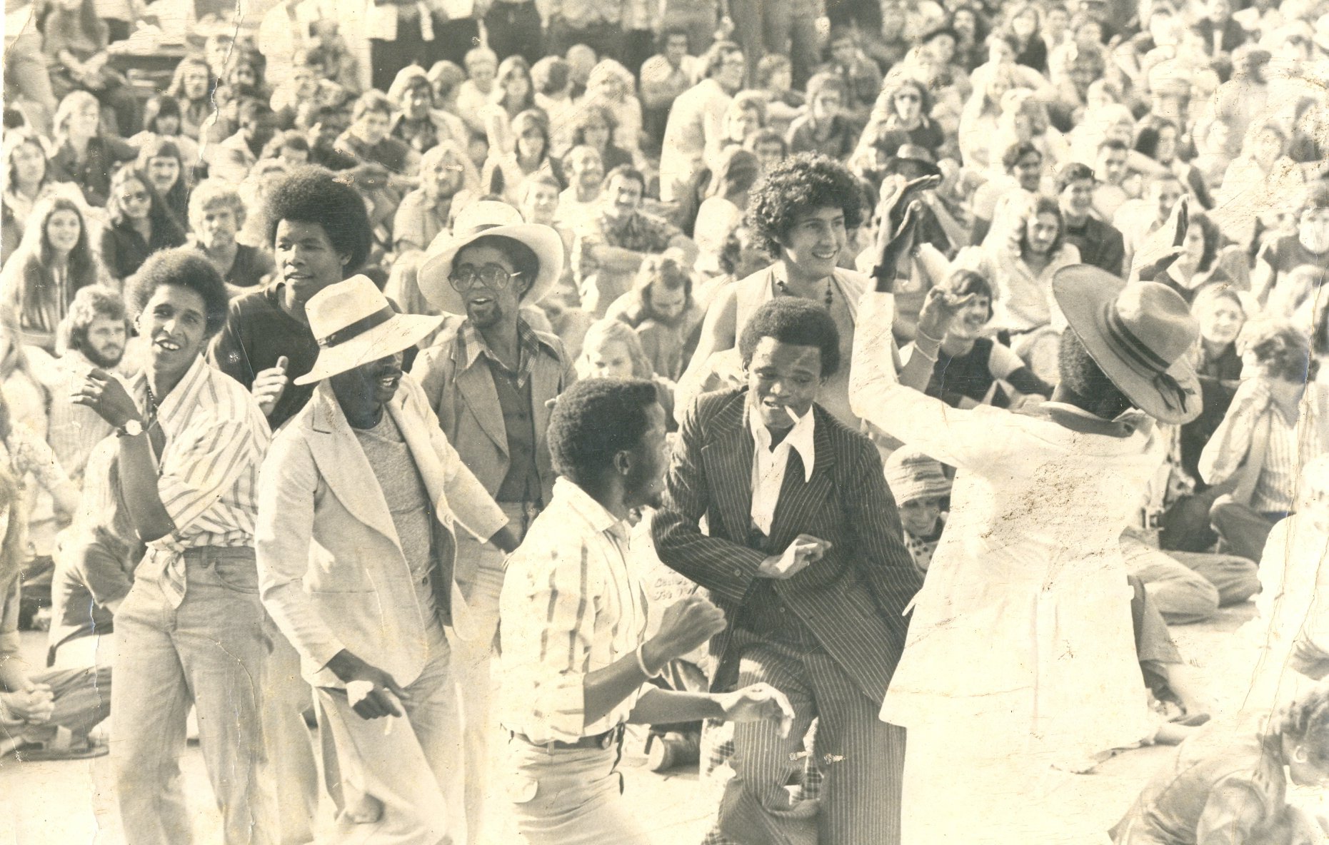 L'image représente une scène vibrante de fête en plein air, probablement lors d'un concert ou d'un festival. Au premier plan, un groupe de personnes danse joyeusement, exprimant de l'énergie et de la convivialité. Les danseurs, de différentes origines, portent des vêtements variés, allant des costumes élégants à des tenues plus décontractées. En arrière-plan, une foule dense assiste à la scène, créant une ambiance de camaraderie. Les visages sont souriants, captivés par la musique et l'atmosphère festive. Le tout suggère une célébration de la vie et de la culture, où la danse et la communion entre les gens sont à l'honneur. L'éclairage semble naturel, évoquant une belle journée ensoleillée.