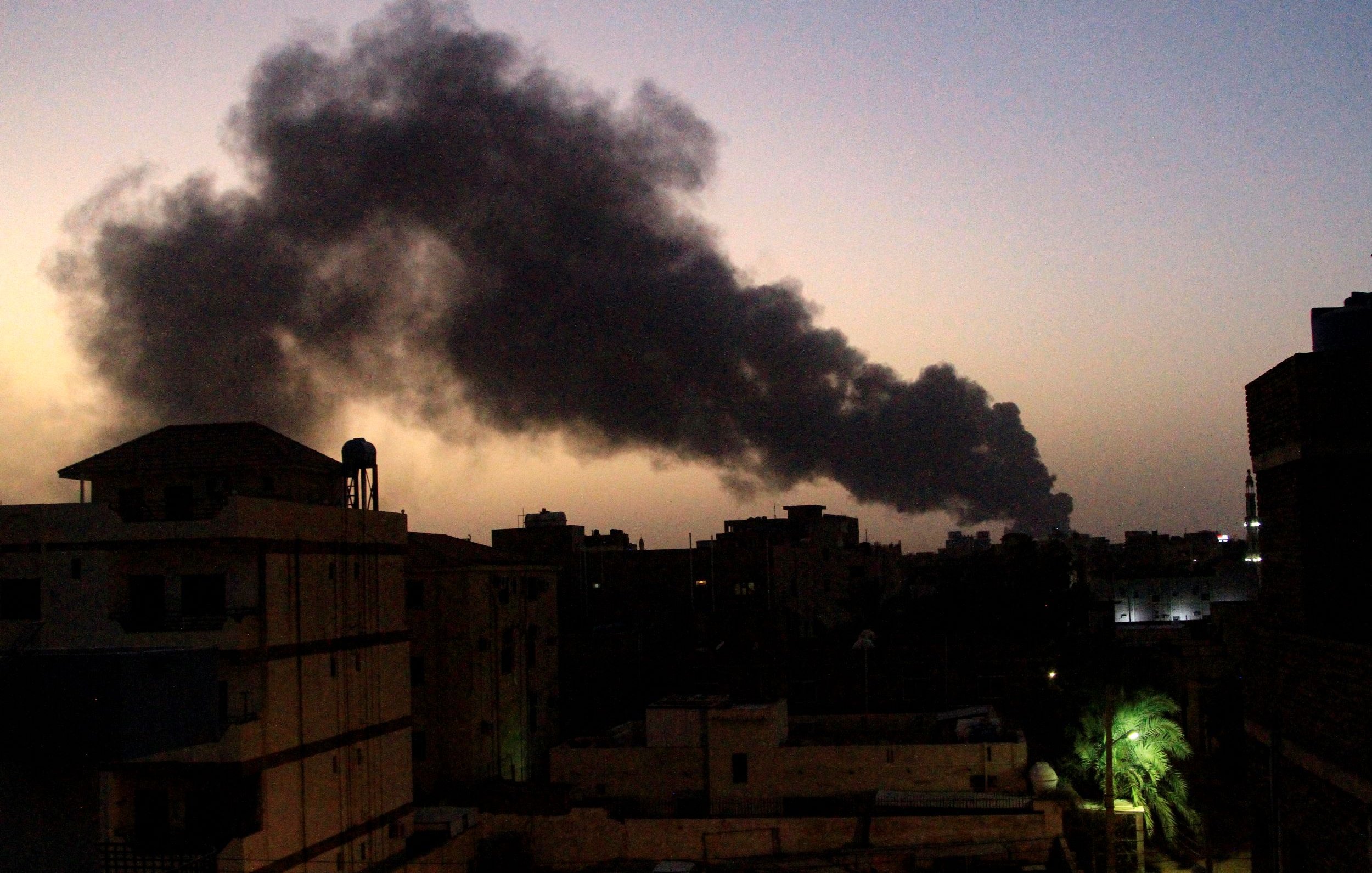 L'image montre un paysage urbain au crépuscule, où le ciel est teinté de nuances de bleu et d'orange. En arrière-plan, une épaisse colonne de fumée noire s'élève, suggérant une source d'incendie. Les silhouettes de bâtiments se dessinent en avant-plan, avec des structures variées et quelques lumières allumées. À droite, on aperçoit un palmier, ajoutant une touche de verdure à ce paysage sombre et chargé d'une ambiance d'urgence.