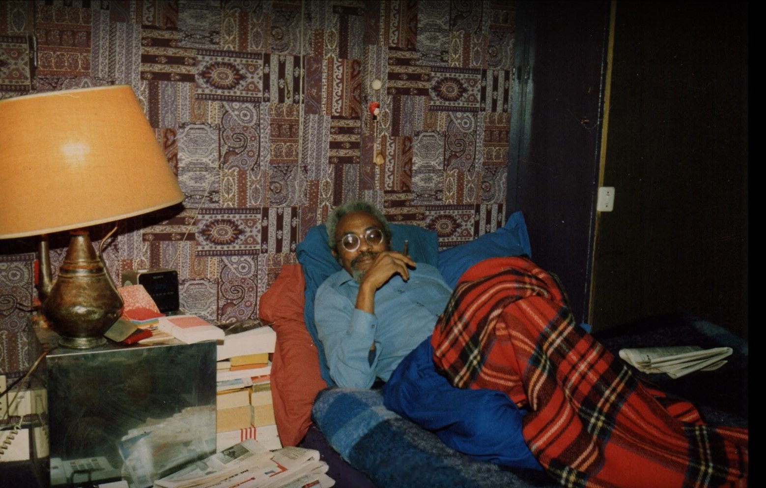 L'image montre un homme allongé sur un lit, recouvert d'une couverture à carreaux rouges et noirs. Il est vêtu d'une chemise bleue et porte des lunettes. L'homme semble pensif, avec un léger sourire. En arrière-plan, on distingue un mur tapissé d'un motif coloré et complexe. À côté du lit, une lampe de table en métal émet une lumière douce. Il y a également des piles de livres et de journaux sur une table à proximité, ce qui donne une impression de confort et d'intimité dans la pièce.