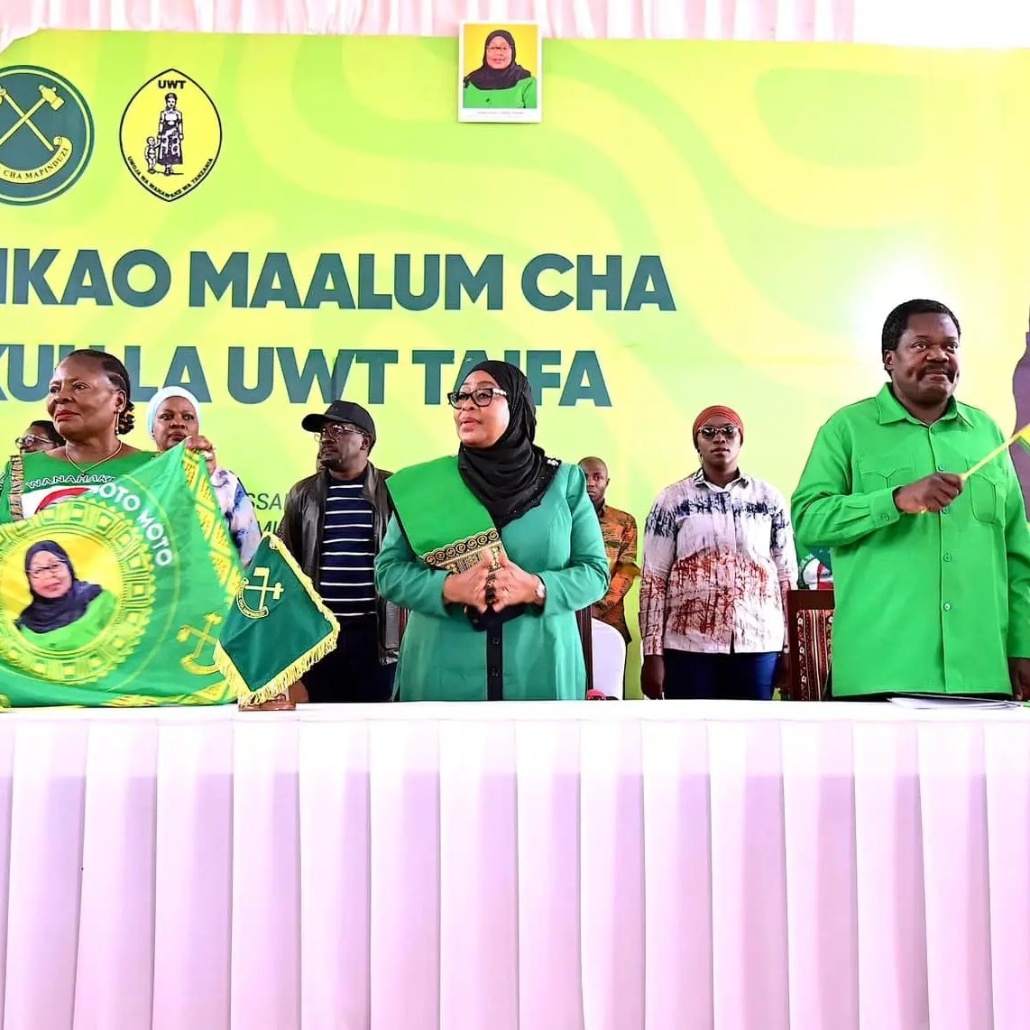 L'image montre un événement politique avec plusieurs personnes debout derrière une table, vêtues de chemises vertes. Au fond, on peut voir une grande bannière avec un portrait d'une femme souriante, probablement une dirigeante. Les participants tiennent des drapeaux ou des banderoles, souvent en vert et jaune, qui portent des symboles liés à leur organisation. L'atmosphère semble festive et engagée, avec des membres du public en arrière-plan qui écoutent attentivement. La réunion semble évoquer des thèmes de solidarité et de mobilisation.