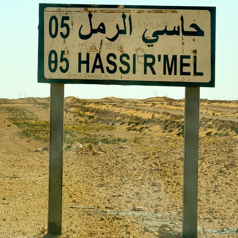 L'image montre une route désertique dans un paysage aride. À droite, un grand panneau de signalisation indique "Hassi R'Mel" en arabe et en français, avec le numéro 05. En arrière-plan, on aperçoit des structures industrielles, dont une cheminée qui dégage de la fumée, suggérant l'activité d'extraction de ressources. L'environnement est principalement constitué de terres sèches et peu de végétation, illustrant une atmosphère isolée et stérile. Le ciel est clair, et la chaleur du jour est palpable.