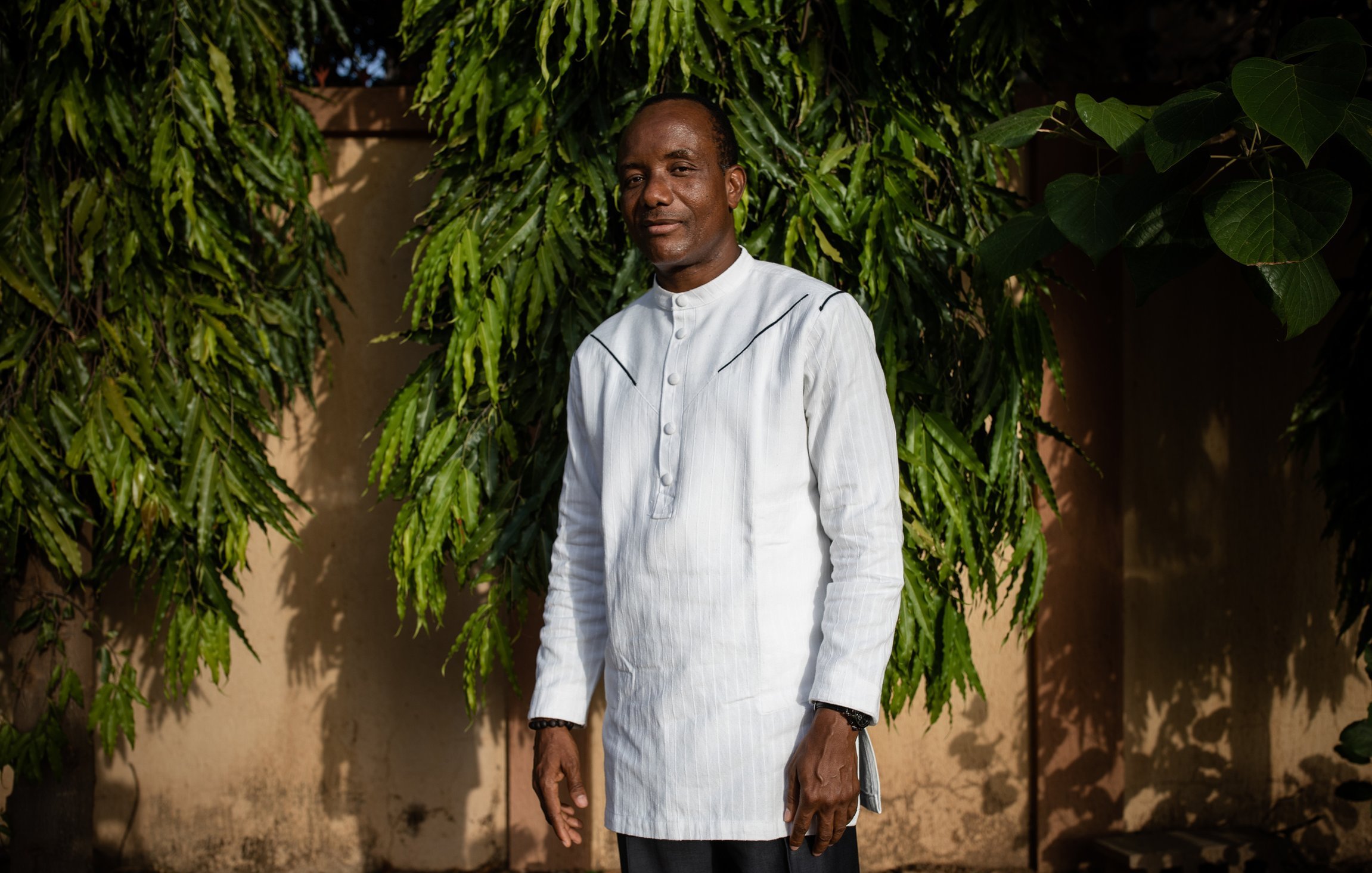 L'image montre un homme debout devant un arrière-plan de feuillage vert dense. Il porte une tunique blanche à col montant et un pantalon sombre. Son sourire dégage une attitude amicale et confiante. Les arbres autour de lui ajoutent une ambiance naturelle, avec des branches feuillues qui encadrent sa silhouette. Le fond est constitué de murs dans des tons nude, apportant une touche chaleureuse à la scène. La lumière naturelle semble douce, créant une atmosphère sereine.