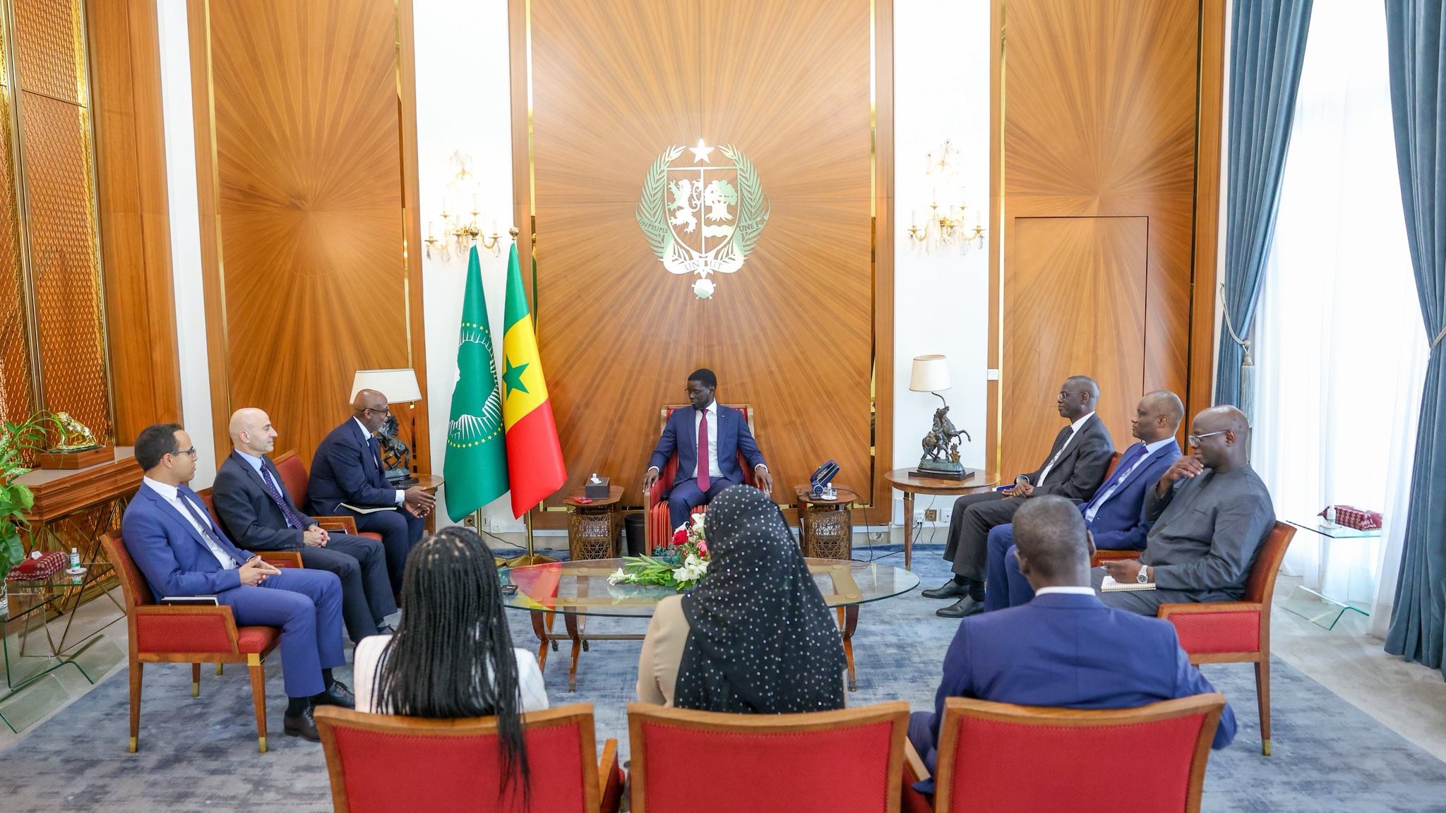 L'image montre une réunion dans une salle élégante. Au centre, un homme assis porte un costume et semble être à la tête de la discussion. Autour de lui, plusieurs personnes assisent dans des fauteuils confortables. À l'arrière-plan, on peut voir les drapeaux du Cameroun et du Sénégal. La pièce est lumineuse, avec des rideaux drapés et un décor raffiné, donnant une atmosphère formelle et professionnelle à cette rencontre. Les participants semblent engagés dans une conversation sérieuse.