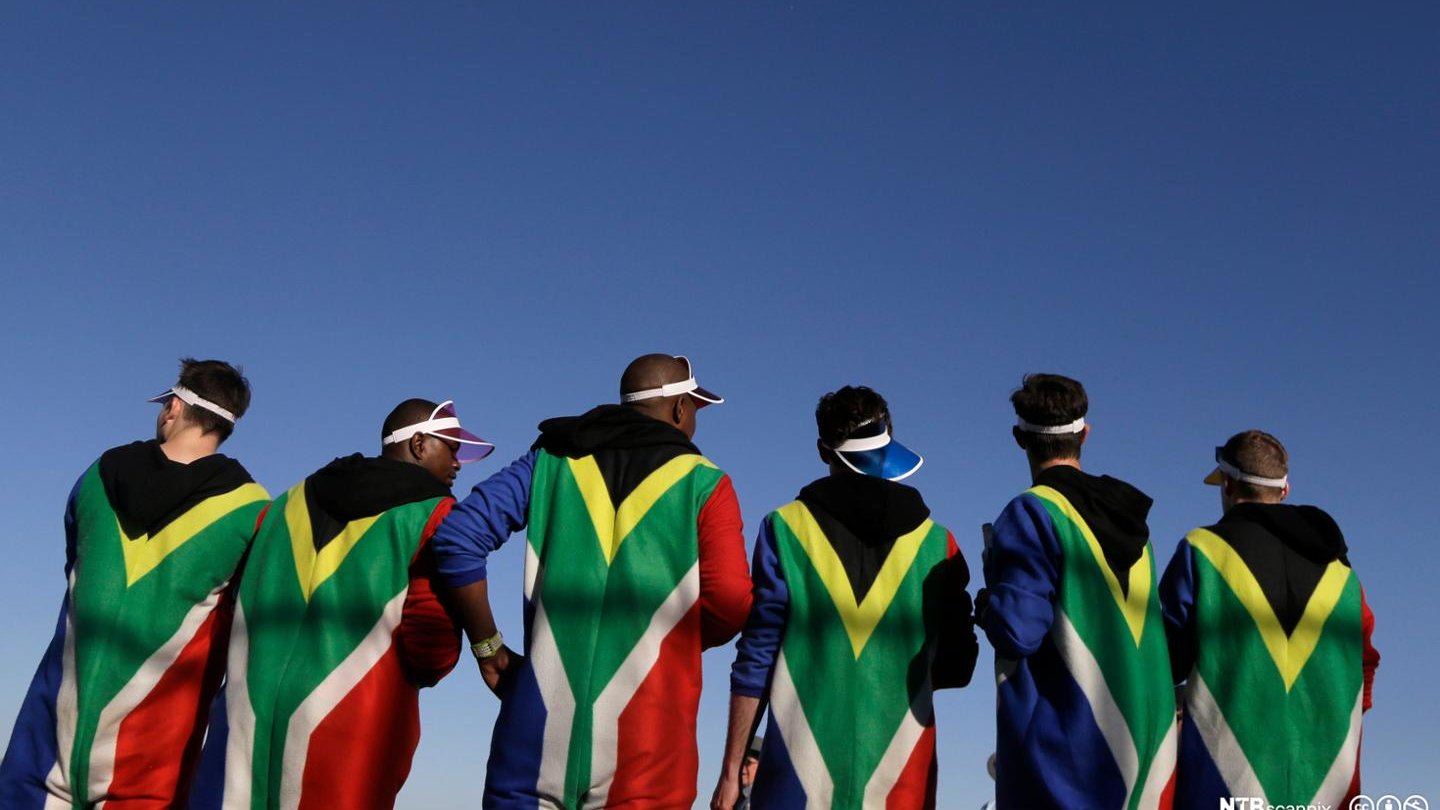 L'image montre un groupe de personnes de dos, habillées dans des capes aux couleurs du drapeau sud-africain, dominant les tons vert, noir, jaune et rouge. Leurs silhouettes sont nettement visibles contre un ciel bleu clair. Les individus semblent détendus et peuvent être en train de profiter d'un événement ou d'une célébration. Certains portent des visières sur la tête, ajoutant une touche festive à leur tenue. L'ensemble dégage une ambiance dynamique et communautaire, soulignant la fierté et l'identité culturelle.