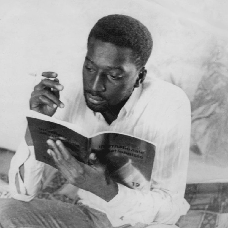 L'image montre un homme assis, plongé dans la lecture d'un livre. Il a la peau foncée et porte une chemise blanche. Dans sa main droite, il tient une cigarette. L'ambiance de la scène est calme, suggérant un moment de réflexion ou d'étude. L'arrière-plan évoque un espace intime, avec des motifs sur le sol qui peuvent donner une impression de confort. L'homme semble absorbé par sa lecture, dégageant une concentration et une sérénité.