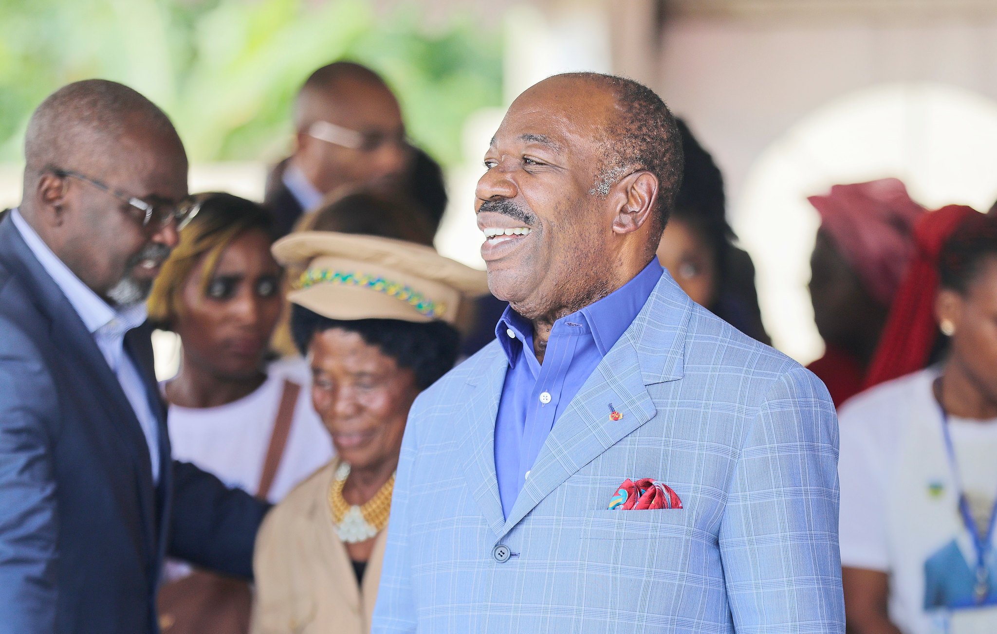 L'image montre un groupe de personnes en train d'interagir dans un cadre convivial. Au centre, un homme, portant un costume bleu clair et une chemise bleue, sourit chaleureusement, affichant une attitude amicale et accessible. On note également des personnes en arrière-plan, certaines portant des vêtements colorés, qui semblent engagées dans une conversation ou une activité sociale. L'ambiance globale de l'image est joyeuse et accueillante, suggérant un moment de célébration ou de rencontre.
