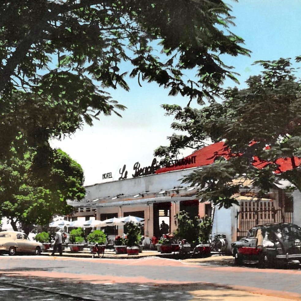L'image montre une scène d'un restaurant, probablement située dans un quartier ensoleillé. À l'avant, on aperçoit une route bordée d'arbres dont les feuilles offrent un peu d'ombre. Le bâtiment principal, assez spacieux, possède un toit rouge et est marqué par l'inscription "Le Restaurant" en lettres blanches. Plusieurs voitures anciennes, de différentes couleurs, sont garées près de l'établissement. Des personnes flânent autour, créant une atmosphère de vie et de convivialité. En arrière-plan, le ciel est bleu, ce qui ajoute une touche de chaleur et de clarté à l'ensemble de l'image.