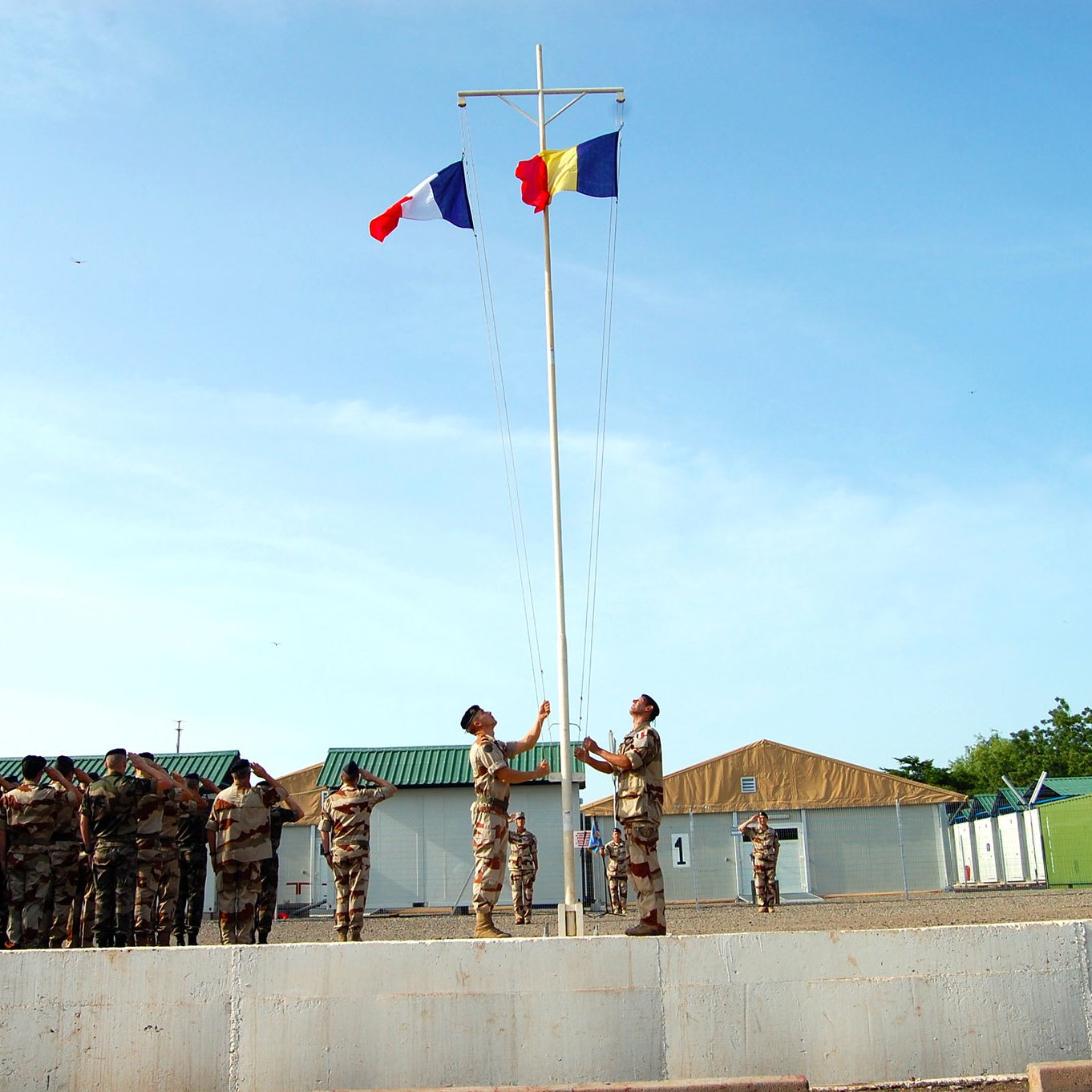 Cette image montre une cérémonie de lever de drapeaux dans un environnement militaire. Au premier plan, deux soldats se tiennent près d'un mât, où ils sont en train de hisser les drapeaux français et roumain. Ils sont parfaitement alignés et semblent respectueux. En arrière-plan, un groupe de soldats en uniforme assiste à la cérémonie, formant une ligne. L'environnement est clair et ensoleillé, avec des bâtiments militaires simples et une légère brise qui pourrait faire flotter les drapeaux. L'atmosphère est solennelle et empreinte de respect.