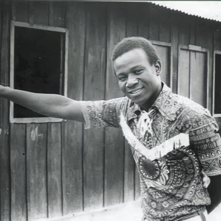 L'image montre un homme debout à côté d'une maison en bois. Il porte une chemise colorée au motif complexe, avec des nuances de marron et de beige. Son sourire est amical et engageant. L'homme pointe une main vers un mur en bois, suggérant une direction ou attirant l'attention sur quelque chose. En arrière-plan, on aperçoit des fenêtres sombres qui ajoutent une dimension de profondeur à la scène. L'atmosphère semble à la fois chaleureuse et authentique, évoquant un moment de convivialité.