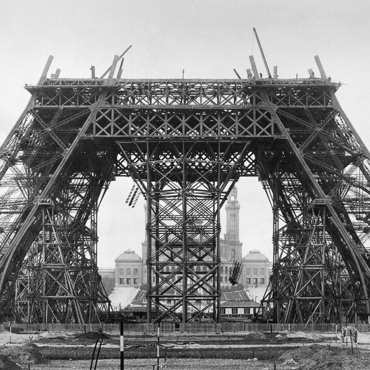 Cette image montre une phase de construction de la célèbre Tour Eiffel à Paris. On peut voir une structure métallique imposante, caractérisée par des poutres en fer entrecroisées. La tour, encore incomplète, présente une base large et stable avec des arcades qui s'élèvent vers le ciel. À l'arrière-plan, on distingue un bâtiment, peut-être une mairie, qui ajoute un contexte urbain à la scène. L'ambiance globale de l'image est monochrome, ce qui renforce l'aspect historique et donne un aperçu des débuts de cette icône de l'architecture. Le sol est encore en chantier, avec des débris et des outils, ce qui rappelle le processus de construction en cours.
