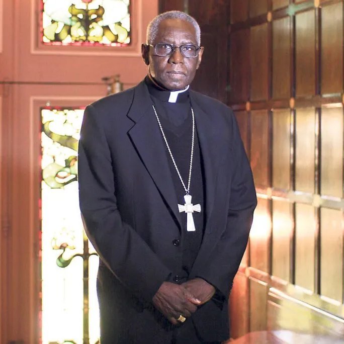 Dans cette image, nous voyons un homme debout dans un environnement qui semble être une pièce élégante, probablement une église ou un bureau religieux. Il porte une tenue noire, typique des membres du clergé, avec une soutane et un col ecclésiastique. Autour de son cou, une grande croix pend, symbolisant son rôle religieux. Le fond est composé de murs en bois foncé et de vitraux colorés, ce qui crée une atmosphère solennelle et respectueuse. Les motifs des vitraux laissent transparaître une lumière douce, ajoutant une dimension spirituelle à l'ensemble de la scène. L'homme a une expression sérieuse, se tenant droit et digne, ce qui évoque l'autorité et la sagesse.