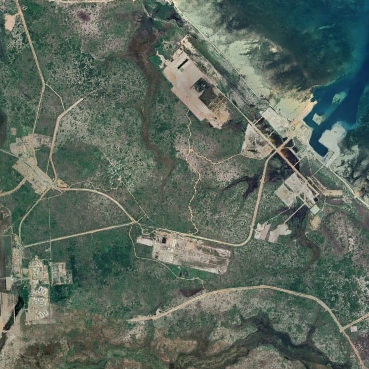 L'image représente une vue aérienne d'une région dont le paysage est marqué par des routes, des structures et des zones déforestées. On peut observer des chemins sinueux qui relient différents points, ainsi que des zones construites qui semblent être des installations industrielles ou militaires. La présence de la mer à droite de l'image ajoute un contraste avec la verdure terrestre, et on peut également voir des étendues d'eau et des zones côtières. Les nuances de couleur vont du vert des zones boisées aux teintes plus claires des infrastructures.