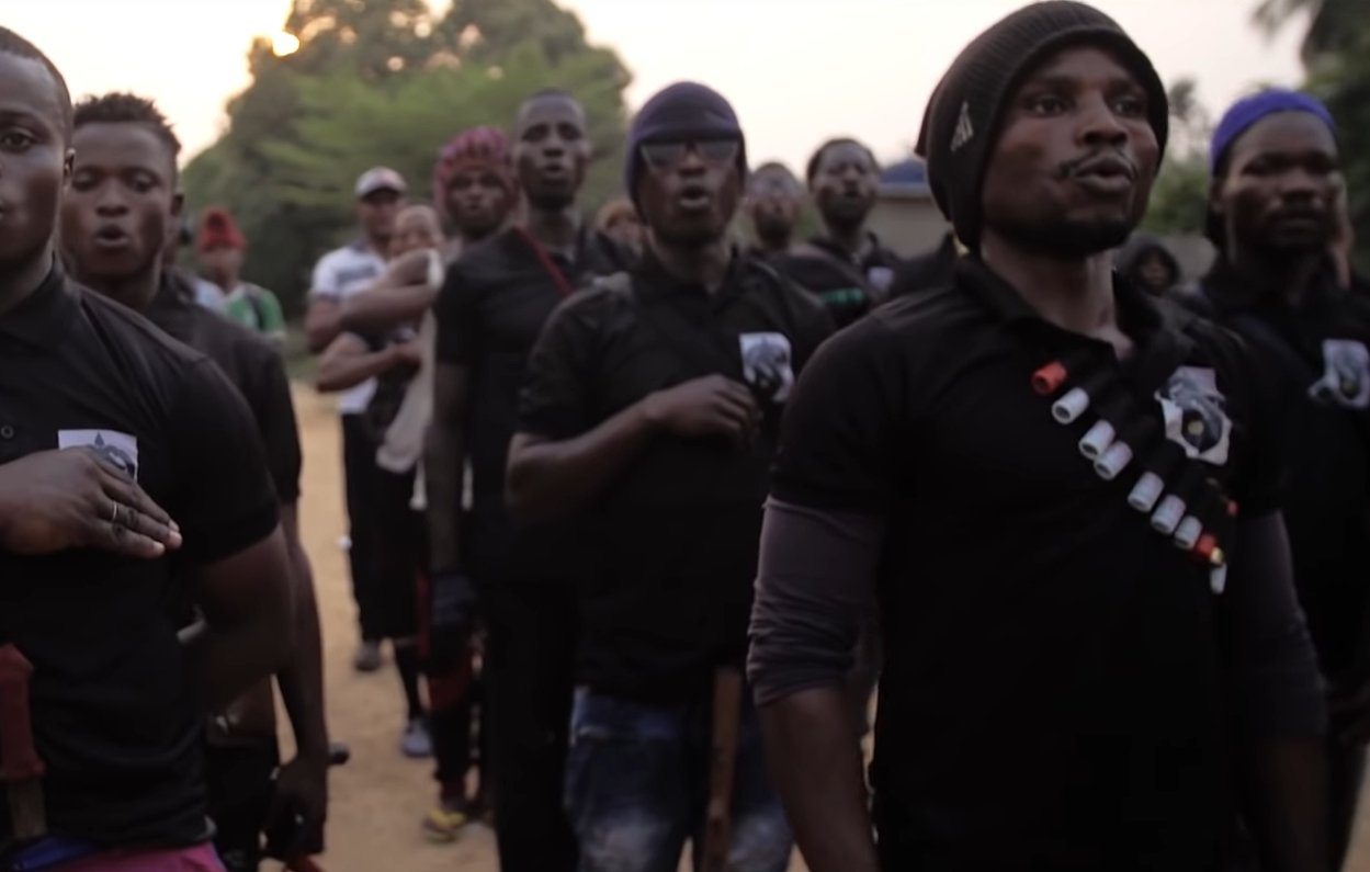 L'image montre un groupe de jeunes hommes debout en formation. Ils portent des t-shirts noirs et semblent participer à une activité collective, probablement un chant ou un cri de ralliement. Certains d'entre eux tiennent des bâtons ou des armes. En arrière-plan, on aperçoit d'autres personnes rassemblées, avec des arbres et un paysage rural. L'atmosphère semble être à la fois solennelle et engagée. La lumière du soir crée une ambiance particulière. 