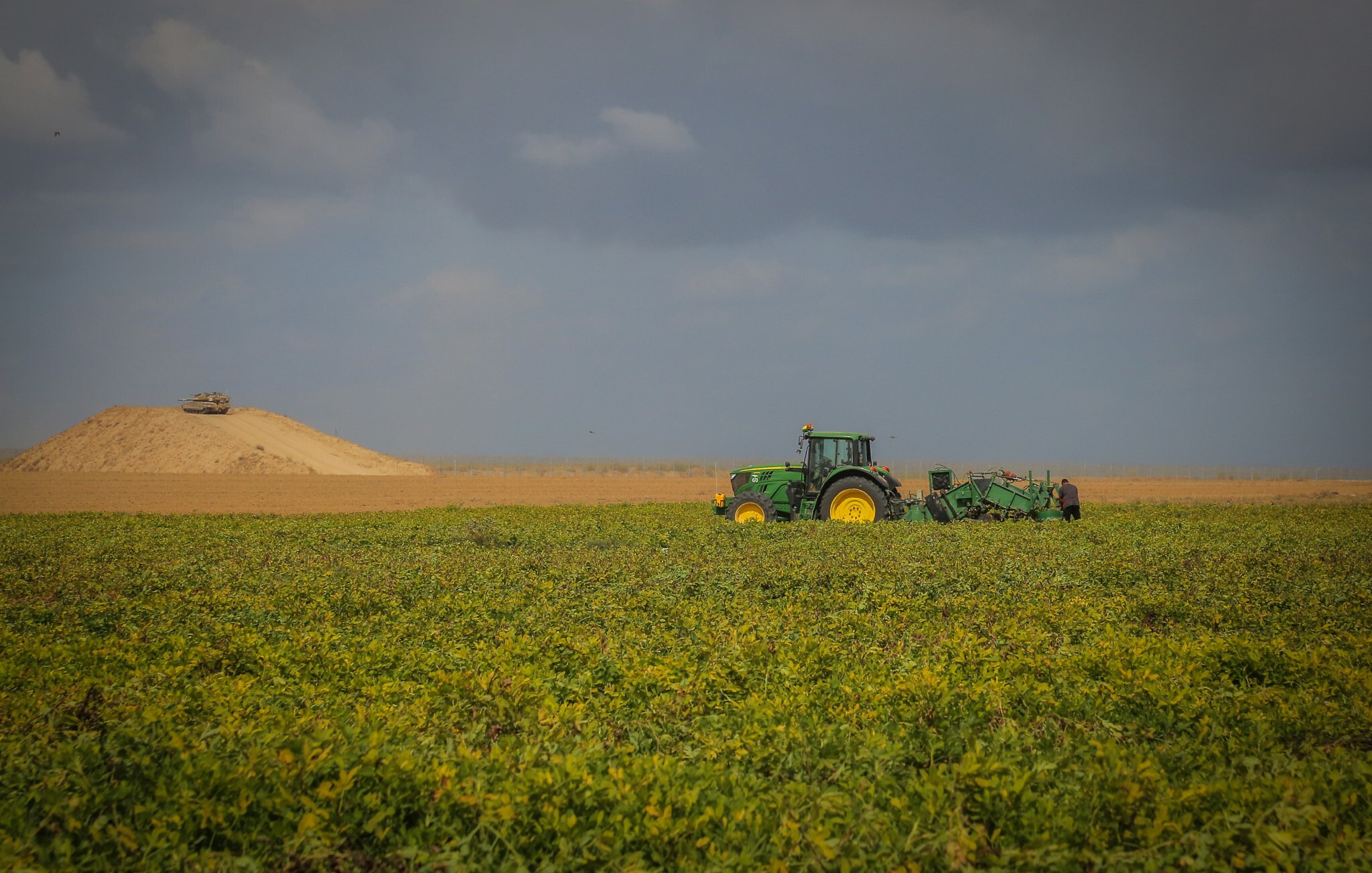 L'image présente un vaste champ de culture où l'on peut voir un tracteur vert, typique des fermes. Le tracteur est engagé dans une activité de labour, préparant le sol pour la plantation. En arrière-plan, on aperçoit une colline de terre argileuse, surmontée d'un véhicule. Le ciel est nuageux, créant une ambiance contrastée avec des teintes de bleu et de gris qui donnent une impression de calme. Les lignes horizontales des cultures et de la colline ajoutent une profondeur visuelle au paysage rural.