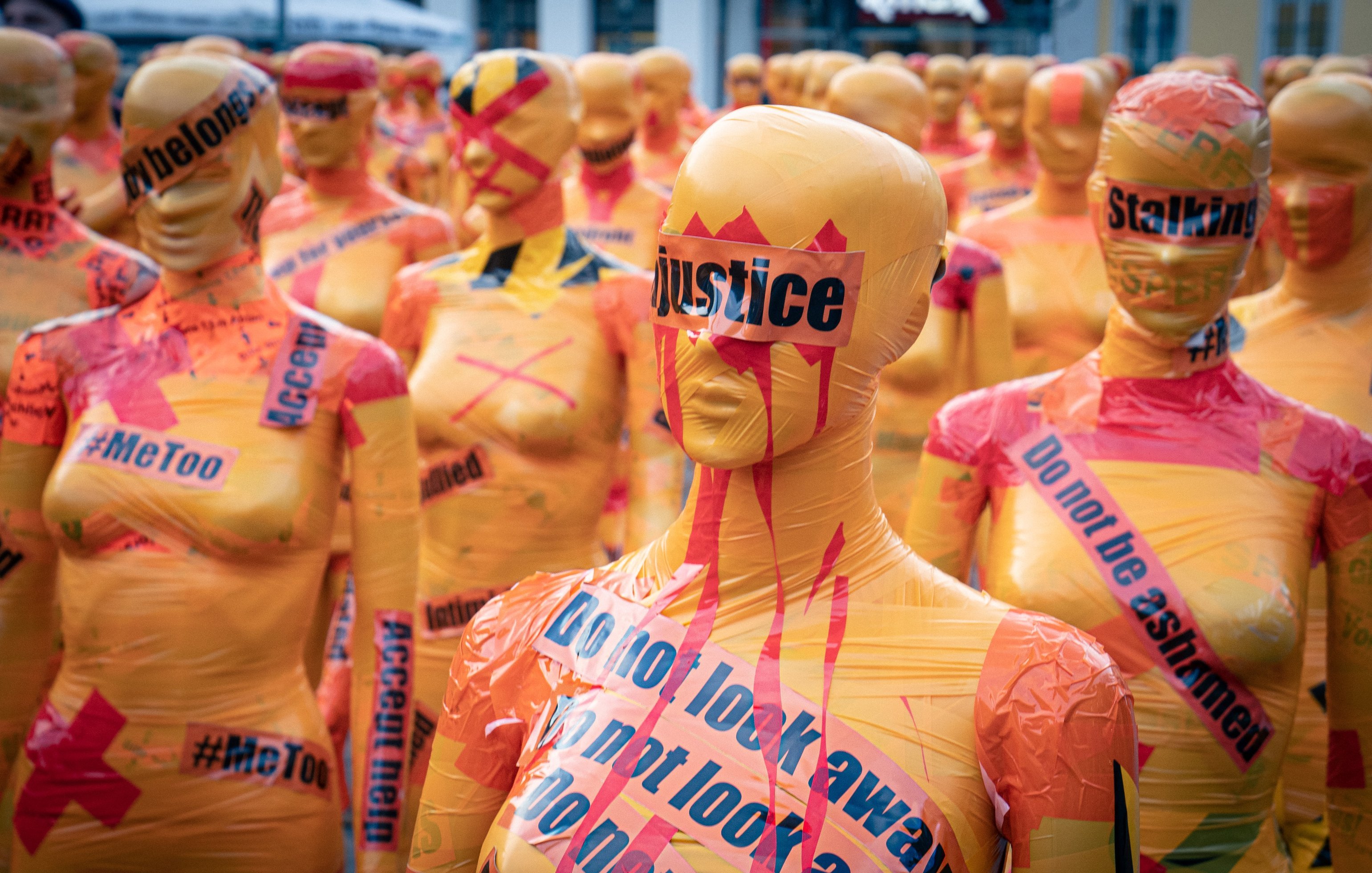 L'image montre un vaste rassemblement de silhouettes humaines couvertes de plastique coloré. Chaque silhouette est peinte en orange vif et ornée de mots et de slogans en noir et en rouge, évoquant des thèmes sociaux et des mouvements de droits civiques comme #MeToo et la justice. Certains mots sont barrés ou soulignés pour accentuer leur impact. L'atmosphère dégage un sentiment fort d'engagement et de protestation, avec ces figures anonymes qui semblent former un mur de revendication. L'ensemble crée une installation artistique saisissante, incitant à la réflexion sur des enjeux sociaux importants.