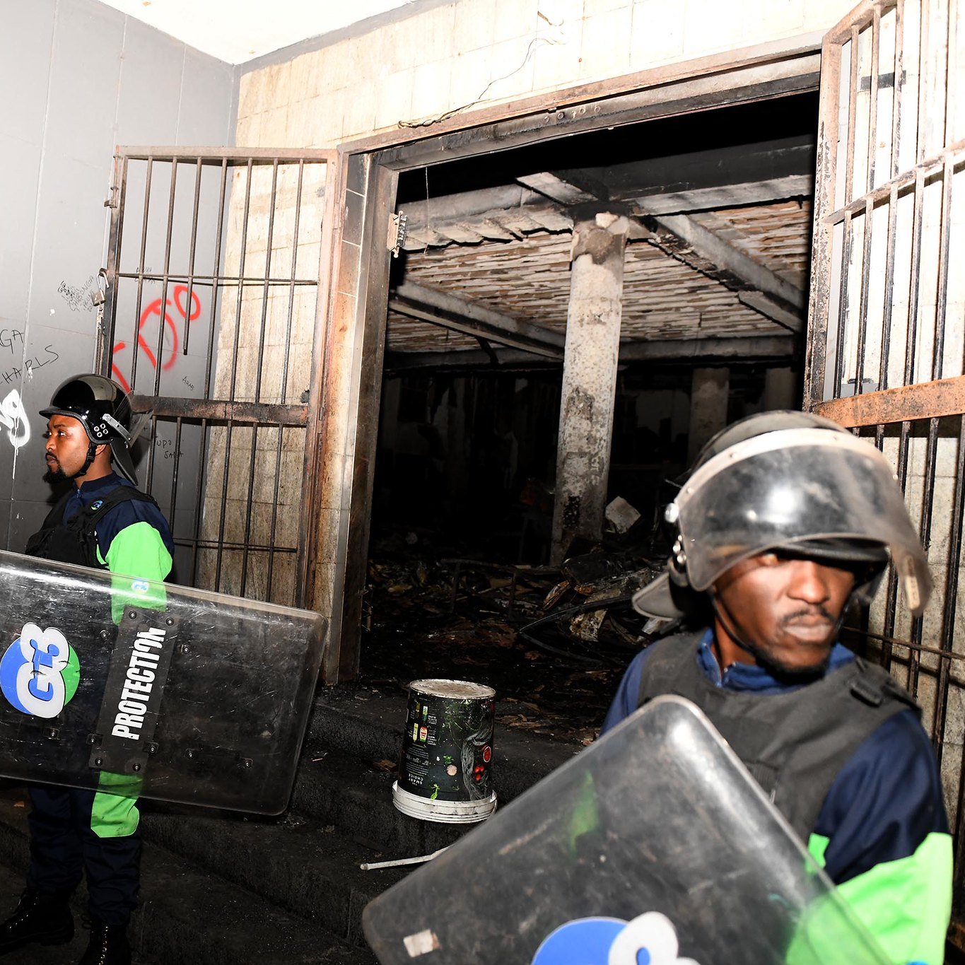 L'image montre une scène de sécurité, probablement après un événement perturbateur. On y voit deux agents de sécurité, vêtus d'un uniforme bleu et vert, portant des casques de protection et tenant des boucliers. Ils se tiennent devant une entrée, à côté de portes métalliques ouvertes. À l'intérieur, on aperçoit un espace sombre avec des traces de dégâts, comme des murs noircis. L'environnement semble chaotique, avec des débris visibles au sol. Des graffitis sont présents sur les murs, ajoutant une touche de désordre à la scène. L'atmosphère générale dégage un sentiment de tension et de vigilance.