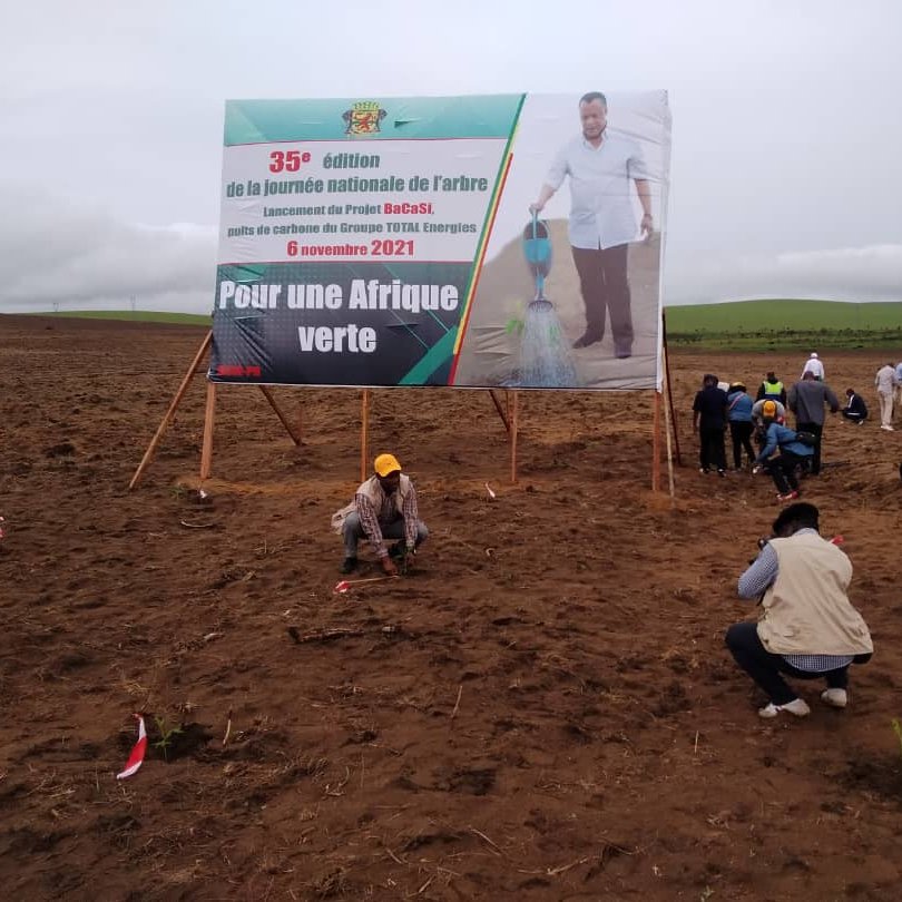 L'image montre un vaste champ de terre nue, où des personnes s'affairent à planter de jeunes arbres. Au centre, un grand panneau affiche le titre "35e édition de la journée nationale de l'arbre" avec une illustration d'un homme en train d'arroser un arbre. La date mentionnée est le 6 novembre 2021, et le slogan "Pour une Afrique verte" est inscrit en bas. Le ciel est nuageux, ce qui suggère un temps frais et peut-être humide, et l'atmosphère semble dynamique, les participants se réunissant pour contribuer à cette initiative de reforestation.