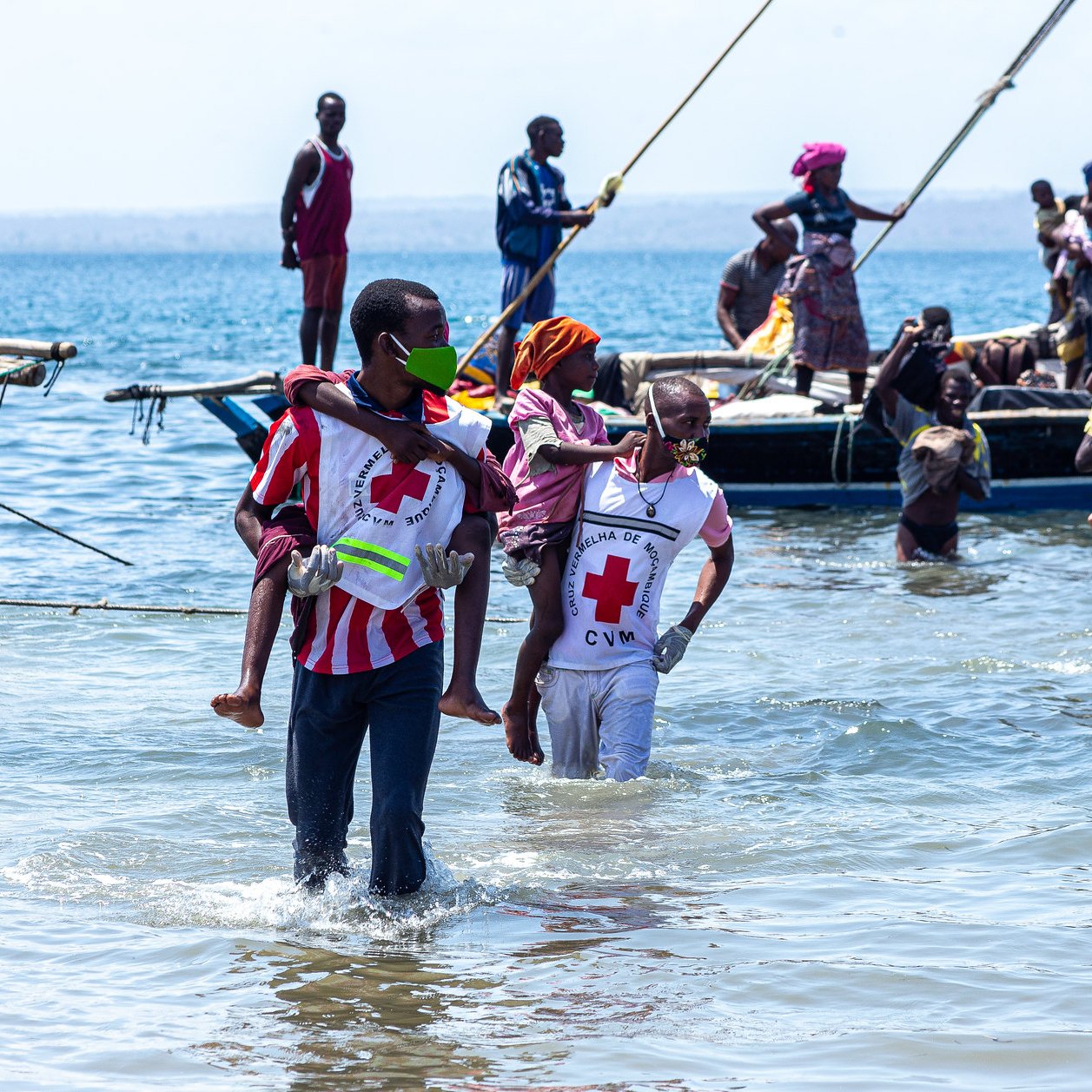 L'image montre une scène animée au bord de la mer. On peut voir plusieurs personnes dans l'eau peu profonds, certaines portant des vêtements colorés. Des hommes et des femmes s'affairent autour de petites embarcations. Certaines personnes transportent des enfants, tandis que d'autres semblent s'entraider. En arrière-plan, il y a une barque avec des passagers qui attendent. L'ambiance semble être pleine de vie et de solidarité, avec un ciel dégagé et une mer calme.