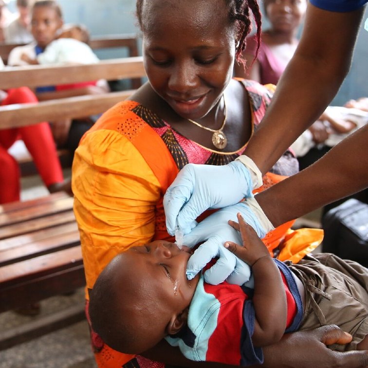 Dans cette image, nous voyons une scène dans une salle d'attente où plusieurs personnes sont assises sur des bancs en bois. Au premier plan, une mère, vêtue d'une robe orange, tient son bébé. Elle semble attentive et douce. Un professionnel de la santé, portant des gants bleus, est en train de donner un soin ou un médicament au bébé. Le bébé, habillé d'un t-shirt bleu et d'un short, a une expression calme. À l'arrière-plan, d'autres femmes et enfants attendent, créant une atmosphère de communauté et de soutien. Les murs sont peints en bleu, ajoutant une ambiance sereine à la scène.